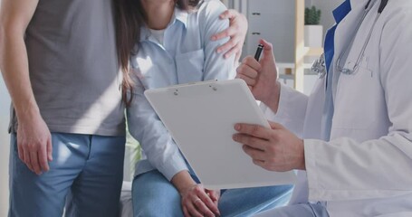 Young couple having fertility treatment. Mid section unrecognizable man hugs and supports woman while listening to clinic doctor with clipboard and pen during health checkup exam appointment visit