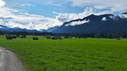 Obraz premium Dirt road is leading to a group of wooden houses in a green field, with mountains and a blue sky with clouds in the background, Garmisch-partenkirchen, Germany