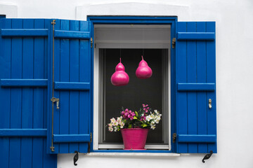 An open window decorated with a pot of flowers and hanging pink squashes. Blue wooden blinds i...