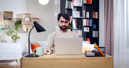Portrait of Arabian male wearing glasses sits in front of laptop screen. Bearded face man works...