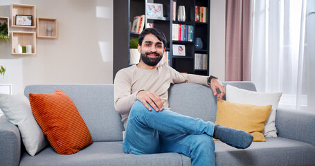 Obraz premium Arabian male sitting on sofa in the middle of room full of different furniture. Handsome bearded face man smiling spends time at own flat.