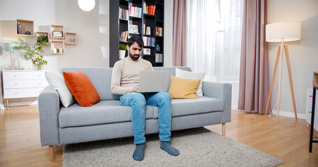Obraz premium Arabian sitting on sofa in the middle of room full of different furniture. Bearded face man orders things online with laptop on knees.