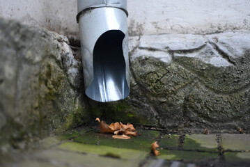 There are wet tiles and autumn leaves under the drainpipe near the wall of the building.