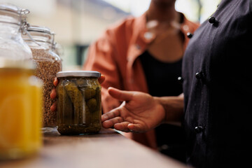 Closeup of african american person with an apron at local market selling fresh eco-friendly produce, advising customer on sustainable food choices. Female seller showing client glass jar of organic