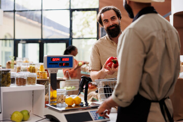 Man with an apron checking the weight of produce using modern scale and selling fresh items to caucasian customer. Local grocery store vendor weighing fruits and vegetables for client.