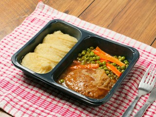 A meal consisting of burger, mashed potatoes and vegetables in plastic tray is served on a red and white checkered tablecloth and wooden table. Simple classic dish for take away. Office lunch.