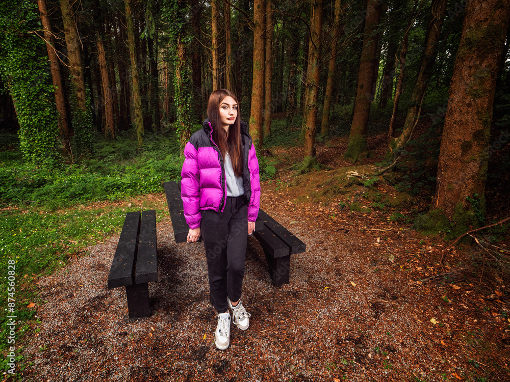 Wall mural Young teenager girl in a dark forest park. Travel and tourism concept. Model is with long dark hair and slim body type wearing colorful jacket and black trousers.