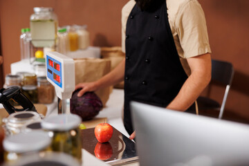 Caucasian store worker in black apron, scans and checks a red apple on weight scale, while he holds a big purple cabbage in his hand. Salesman measuring natural organic produce at cashier desk.
