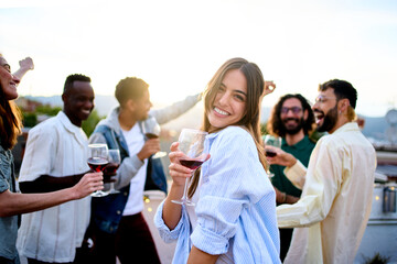 Young Caucasian pretty woman poses coquette holding red wine with friends in background dancing at...
