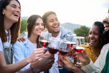 Cheerful group of multiracial friends toasting glasses of red wine together laughing at terrace house outdoors. Cheers of smiling diverse millennial community people having fun at sunset summer party