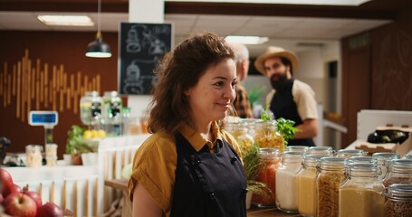 Portrait of smiling woman working in zero waste organic food supermarket, adding pantry staples on shelves. Cheerful employee restocks local grocery store with bulk products in reusable jars