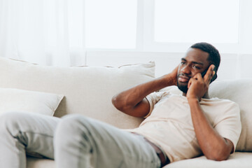 Fototapeta premium Happy African American Man Sitting on Black Sofa, Talking and Typing Messages on Phone in Modern Apartment Enjoying Leisure Time, Relaxing and Connecting with Friends on Social Media