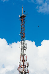 A tall metal tower stands against a bright blue sky, surrounded by puffy white clouds. The tower is a lattice structure, and it is topped with antennas and other equipment.