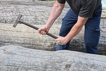 male worker in black blue clothes swings iron hammer in hands over grey wooden log on street