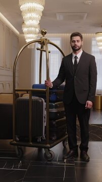 Vertical full portrait of Middle Eastern bellboy in uniform standing with luggage trolley at luxurious hotel lobby