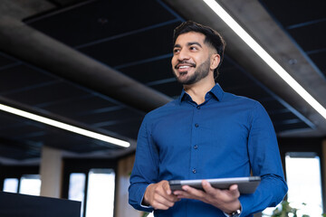 Holding tablet and smiling, businessman standing in modern office environment, copy space