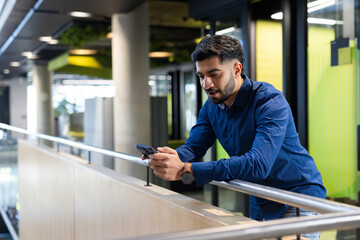 Using smartphone, man leaning on railing in modern office building, copy space