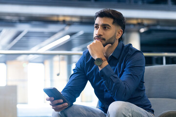 Holding smartphone, thoughtful man sitting in modern office