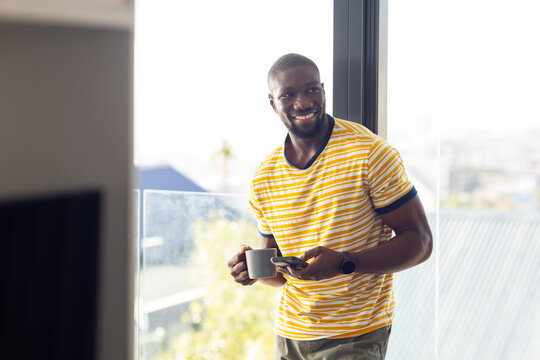 Smiling man holding smartphone and coffee cup, relaxing at home near window