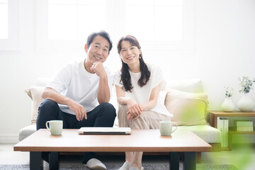 明るい綺麗なリビングでソファに座る　50代夫婦のカメラ目線の広角　緑の前ボケあり