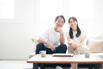 Wide-angle, camera-eye view of a couple in their 50s sitting on a sofa in a bright, clean living room.