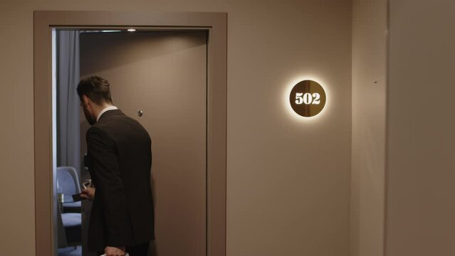Back view medium shot of confident businessman in formalwear with suitcase opening hotel room door with keycard and going in