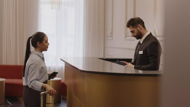 Side view medium shot of young Latin woman with luggage giving her passport to elegant Middle Eastern male receptionist while checking in at luxurious hotel