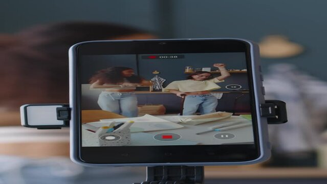Vertical through smartphone display shot of two diverse teenage girls filming dancing video at home using tripod. One of girls holding walking stick because of cerebral palsy