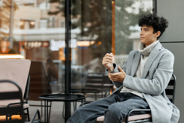 Fashionable young african american man sitting in front of a coffee shop with a cup of coffee in his hand