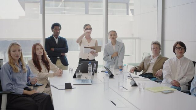 The business meeting features a variety of professionals meeting to discuss collaboration and company strategy in a contemporary office. The group is diverse, engaged, and focused on innovation