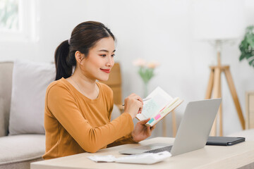 Smiling asian woman working on laptop while sitting in comfortable home with notebook and documents.