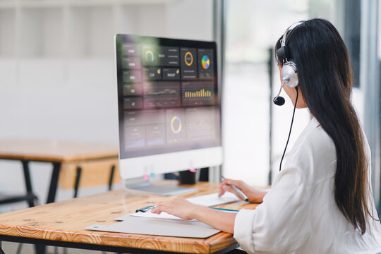 Customer service representative wearing headset analyzes data on computer screen dashboard while working at her desk.
