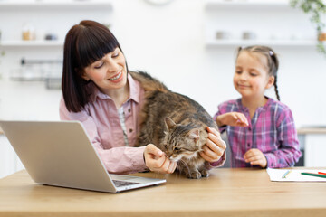 Mother and daughter spending time together while working from home with cat. Woman uses laptop, and girl happily interacts with pet. Cozy home office setup showcasing work balance and family moments.