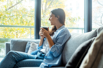 Fototapeta premium Relax and unwind. Young beautiful brunette woman with blissful facial expression alone on the sofa spending good time drinking coffee or tea in cup. Portrait of relaxed woman resting at home.