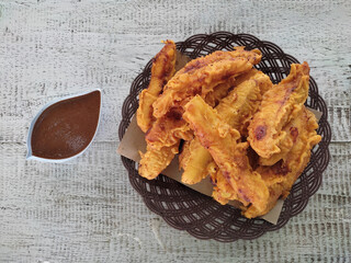 Fried bananas are served on a plastic plate with a woven pattern and placed on a wooden table. Next to it, there is a small bowl containing peanut sauce as a flavor enhancer.