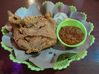 A piece of crispy fried chicken breast, two slices of cucumber, and a small bowl of sambal. They are served on a plastic plate lined with parchment paper.