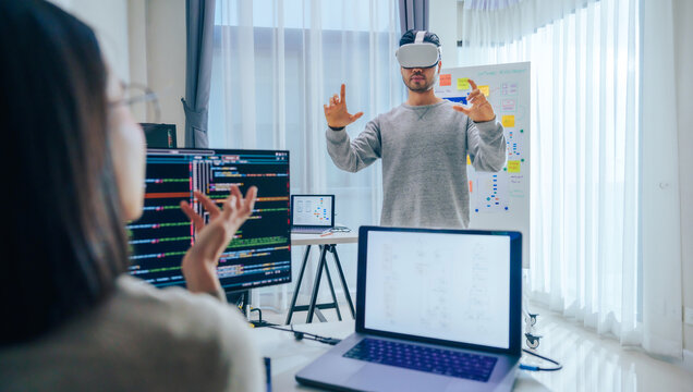 Asian software developer wearing a virtual reality headset works on a VR project, with a colleague focused on her computer