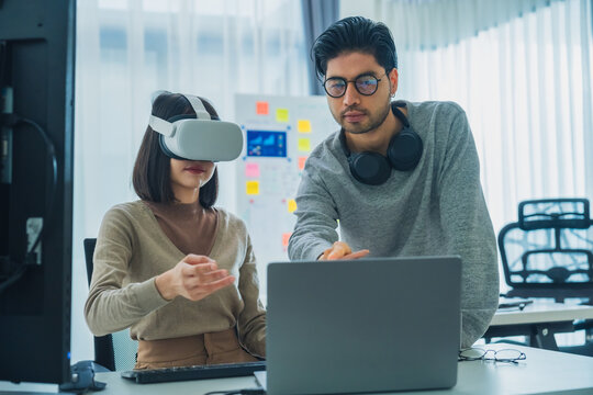 Asian software developer wearing a virtual reality headset works on a VR project, with a colleague focused on her computer