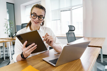 A young professional woman uses a tablet while working in a bright, modern office environment.