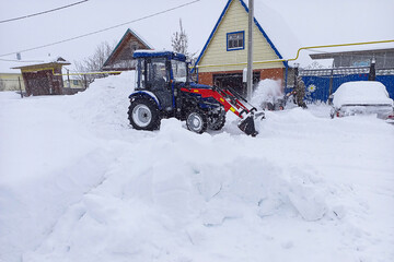 Mini tractor scrapes snow from a covered sidewalk with a bucket while clearing snow on winter. City road services for cleaning, landscaping. Labor mechanization as increase in production efficiency © Po