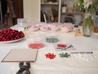 Mosaic pliers, mosaic base, containers with red, green and white glass pieces, a vase with white flowers, a tray with pink zefir and a bowl of ripe cherries on a white surface.