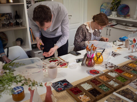 Mosaic Workshop: Horizontal shot of man and woman working at the table with boxes of colourful vitrage glass, pliers, samples of ancient floral patterns, and other tools.