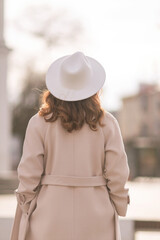 Back view of happy woman wearing hat and coat walking down street on sunny spring day. People, lifestyle, travel and vacations concept