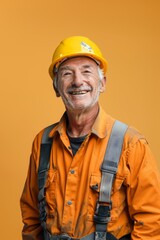 A happy senior construction worker in a hard hat and overalls smiles at the camera against a yellow background