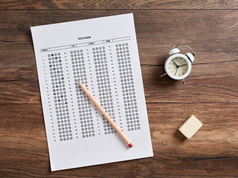 Top view of a test exam paper with alarm clock, pencil and an eraser on a wooden school desk.