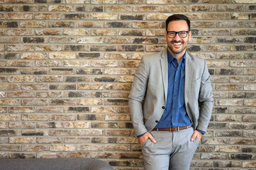 Obraz premium Portrait of handsome CEO with eyeglasses and hands in pockets standing against brick wall in office