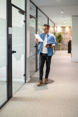 Full length of happy salesman holding laptop and examining sales report while walking in office corridor