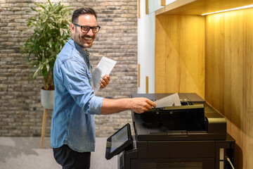 Portrait of male printer smiling and putting sheet of paper into photocopier machine in modern office