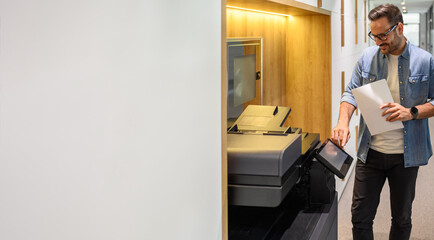 Male assistant in glasses holding financial documents and operating modern computer printer in office