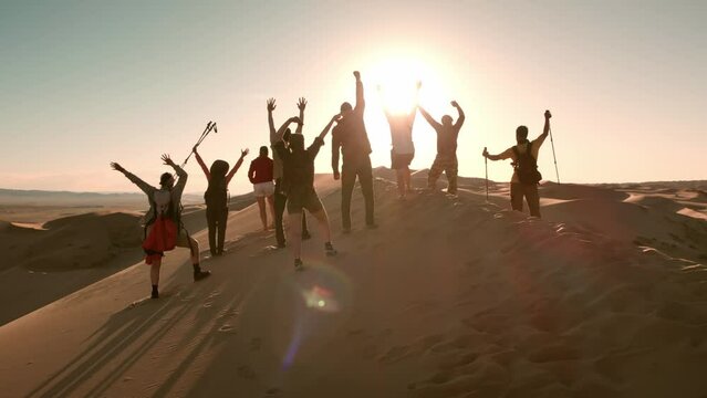 Big group of happy friends, tourists or hikers are standing on dune top and having fun with open arms or winner pose against sunset sky